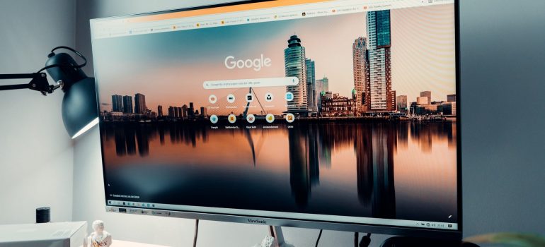 Big computer screen with shown picture of a city and icons of Google, standing on desk and against the grey wall, with a lamp on the left side