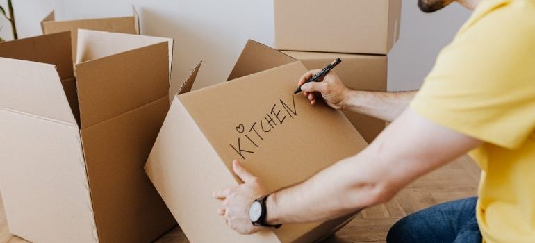 A man labeling a cardboard box "kitchen" while sitting on the floor